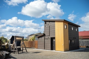 A modern two-story building with a wooden and grey facade, a swing set, and a fenced yard.