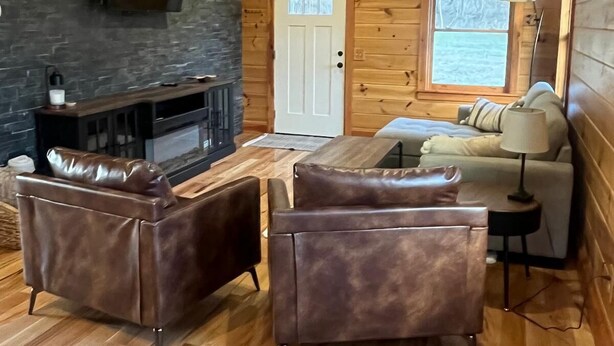 Acogedor interior de cabaña de madera con chimenea, sofás de cuero, televisor y ventilador de techo.
