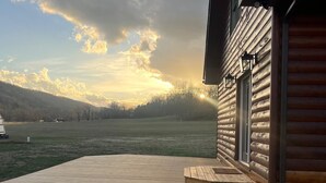 Una terraza de madera que lleva a una cabaña de troncos, con vistas a un campo y colinas al fondo.