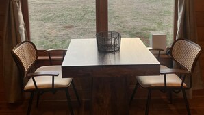 Un comedor con una mesa de madera y dos sillas de mimbre, una cesta sobre la mesa, y vistas a un campo de césped y un granero a través de amplias ventanas.