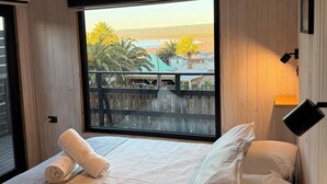 Een slaapkamer met een groot raam, een bed met witte lakens, een houten hoofdbord en een nachtkastje met een afstandsbediening.