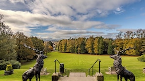 Ein Park mit zwei Hirschfiguren, einer Steintreppe und einer grünen Fläche mit Bäumen.