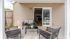 Un patio techado con dos sillones de mimbre negro, una mesa de centro de vidrio y un sofá gris con cojines.