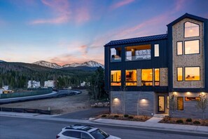 A modern two-story building with large windows and a wooden facade, set against a mountainous backdrop. - VIP Concert Viewing from Private Hot Tub+Walkable+5BR+Sauna+Kids Ski Gear Free (Winter Park)