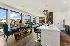 A modern kitchen with a large island, a dining area with a long table and chairs, and large windows offering a view of the outdoors. - VIP Concert Viewing from Private Hot Tub+Walkable+5BR+Sauna+Kids Ski Gear Free (Winter Park)