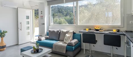 A modern living room with a sofa, coffee table, and bar stools. A large window offers a view of trees and hills.