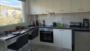 A modern kitchen with white cabinets, a black countertop, a built-in oven, and a window with a view of buildings and trees. - Basil Leaf - Minimal View Retreat (Gonia)