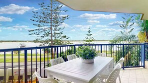 A balcony with a white table and chairs, a potted plant, and a view of the sea and trees.