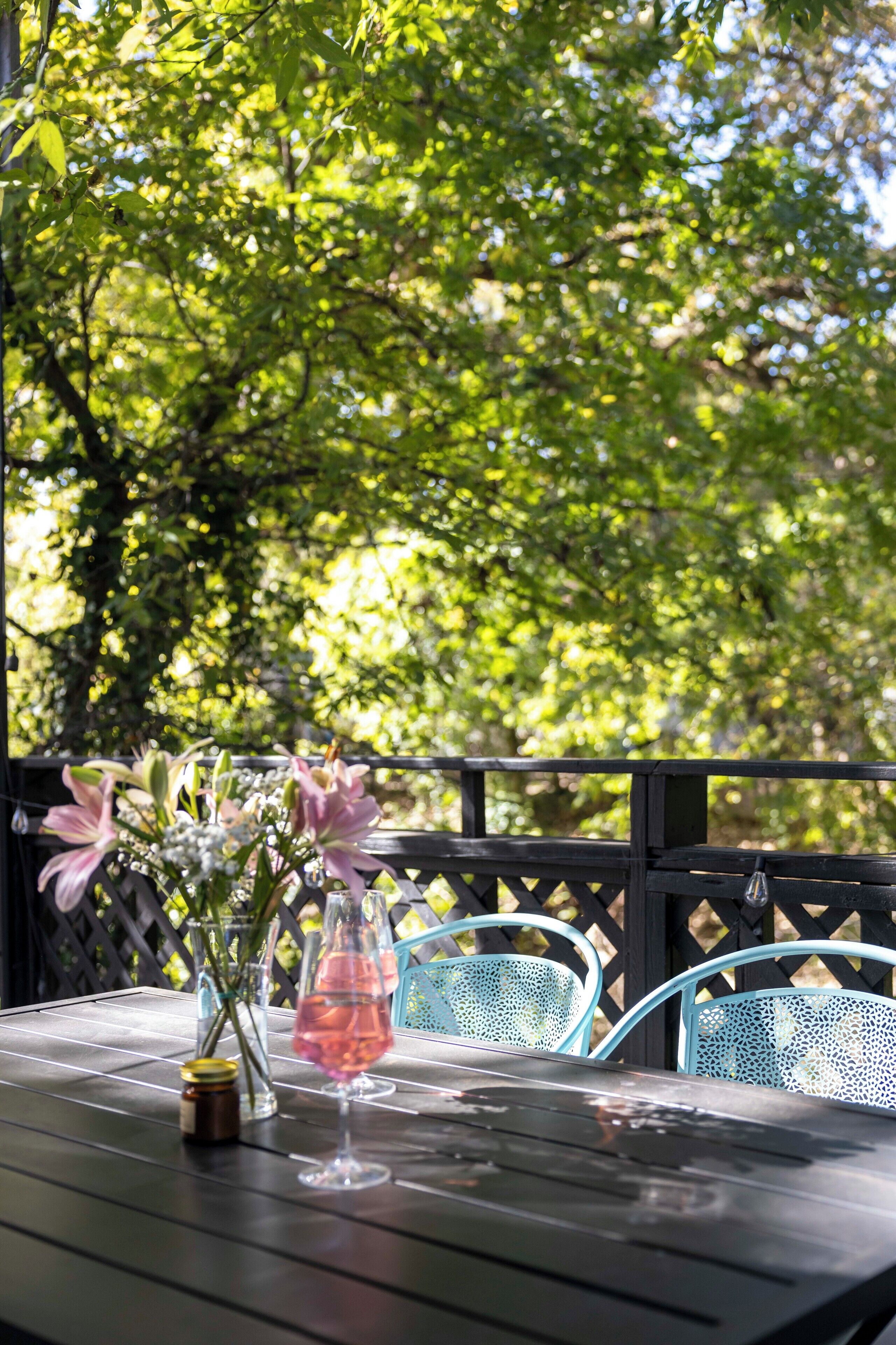 Een tafel met een glas roze drank en een bloevas, voor een achtergrond van groene begroeiing en een houten hek.