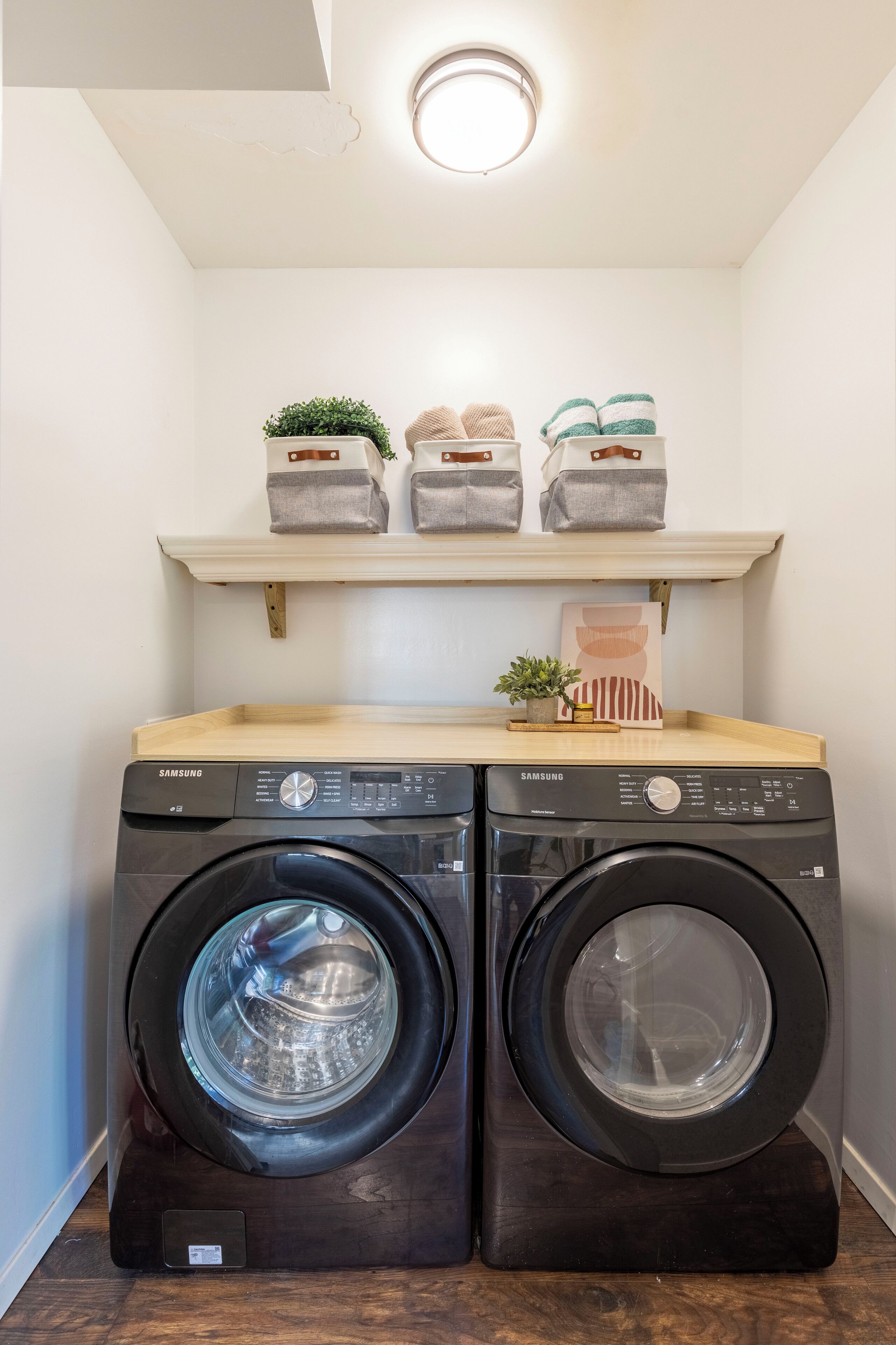 A laundry room with two black Samsung front-loading washers and dryers, a wooden shelf above with decorative items, and a small potted plant.