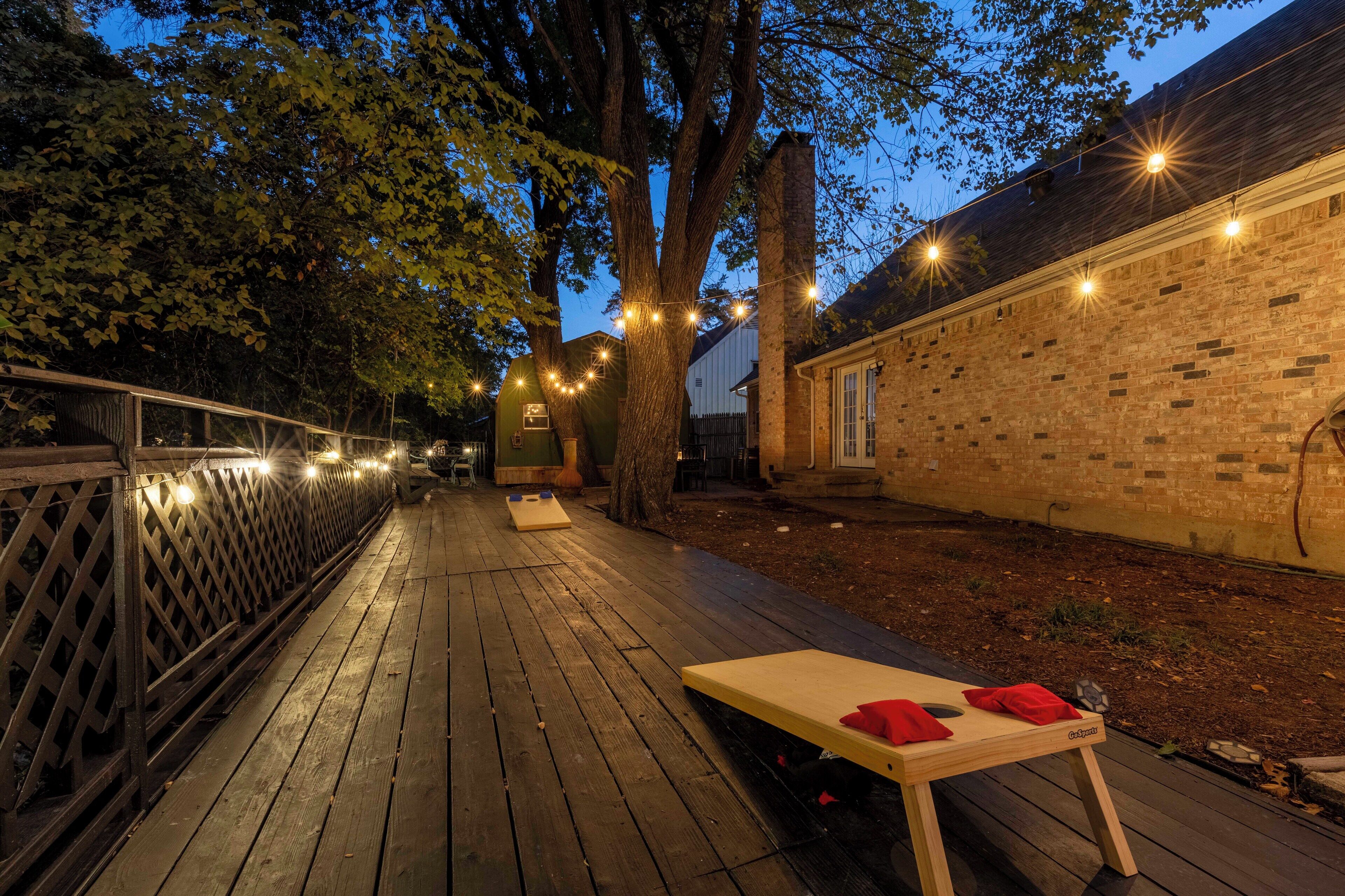 Um deck externo com mesa e cadeiras de madeira, rodeado por árvores e um muro de tijolos.