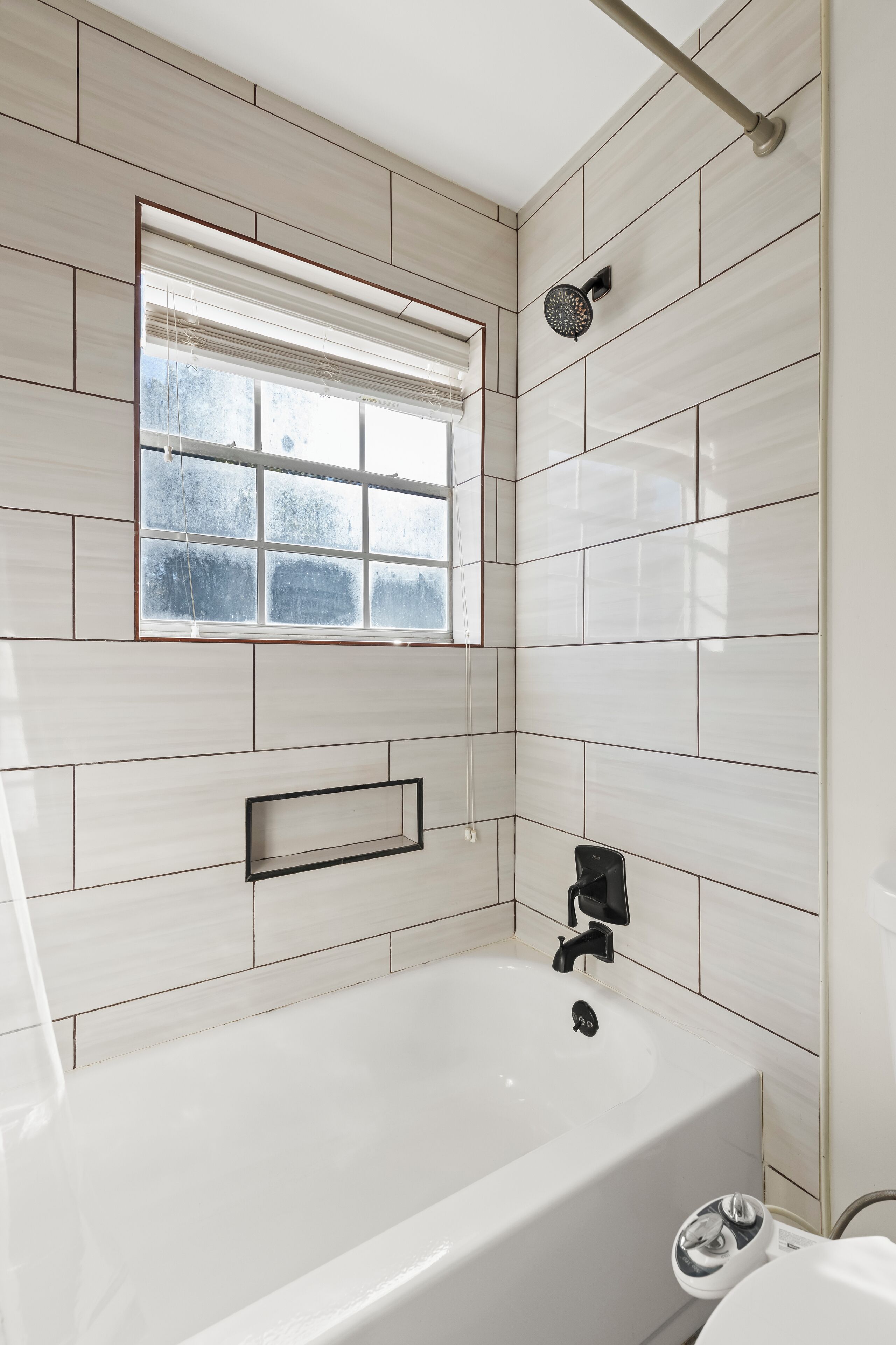 A modern bathroom with a bathtub, a window with blinds, and a showerhead.