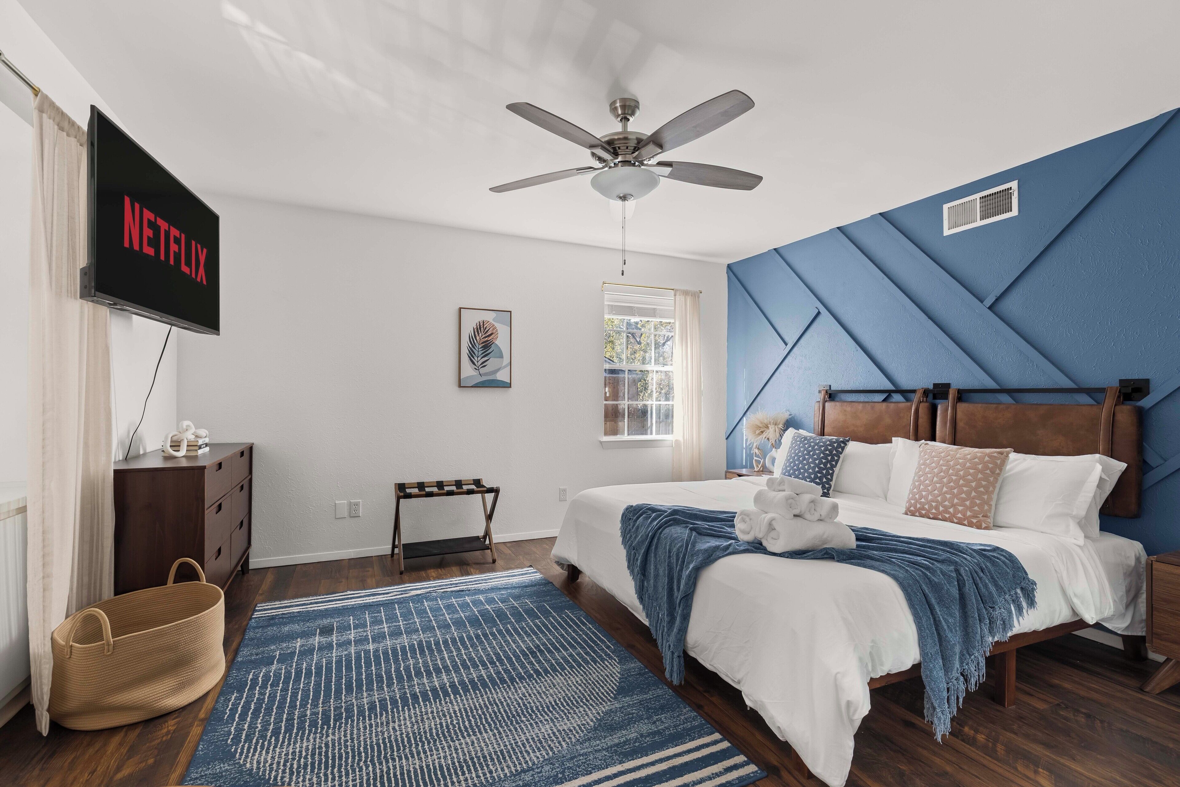 A bedroom with a bed, a ceiling fan, a TV displaying 'NETFLIX', a window, and a wall with a geometric pattern.