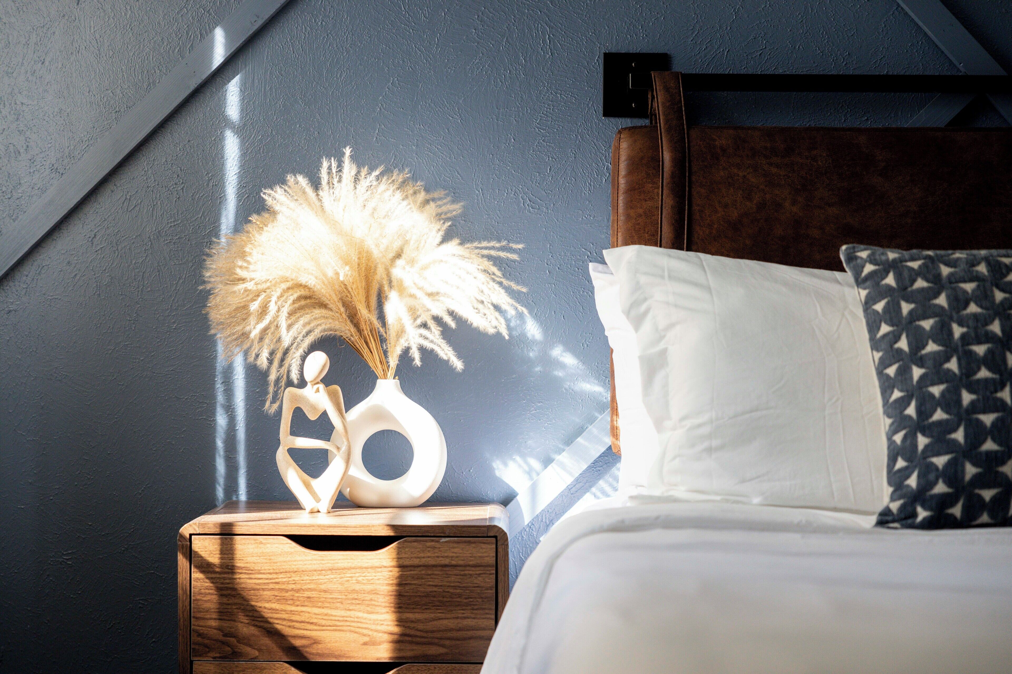 A bedroom with a bed, a nightstand, a decorative vase with pampas grass, and a wall-mounted light switch.