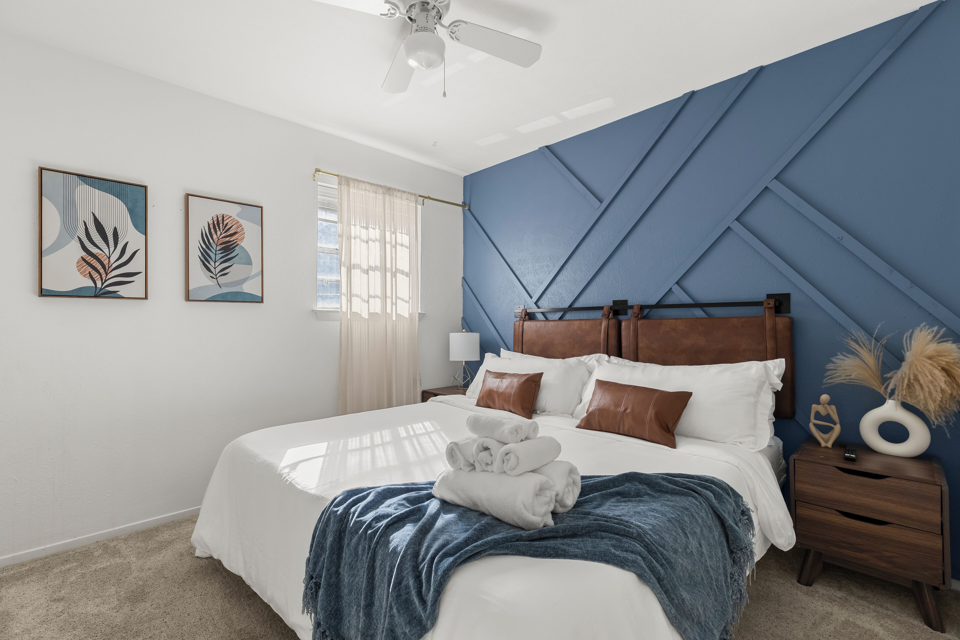 A neatly made bed with rolled towels, a wooden nightstand with a lamp, and two framed artworks on the wall.