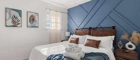 A neatly made bed with rolled towels, a wooden nightstand with a lamp, and two framed artworks on the wall.