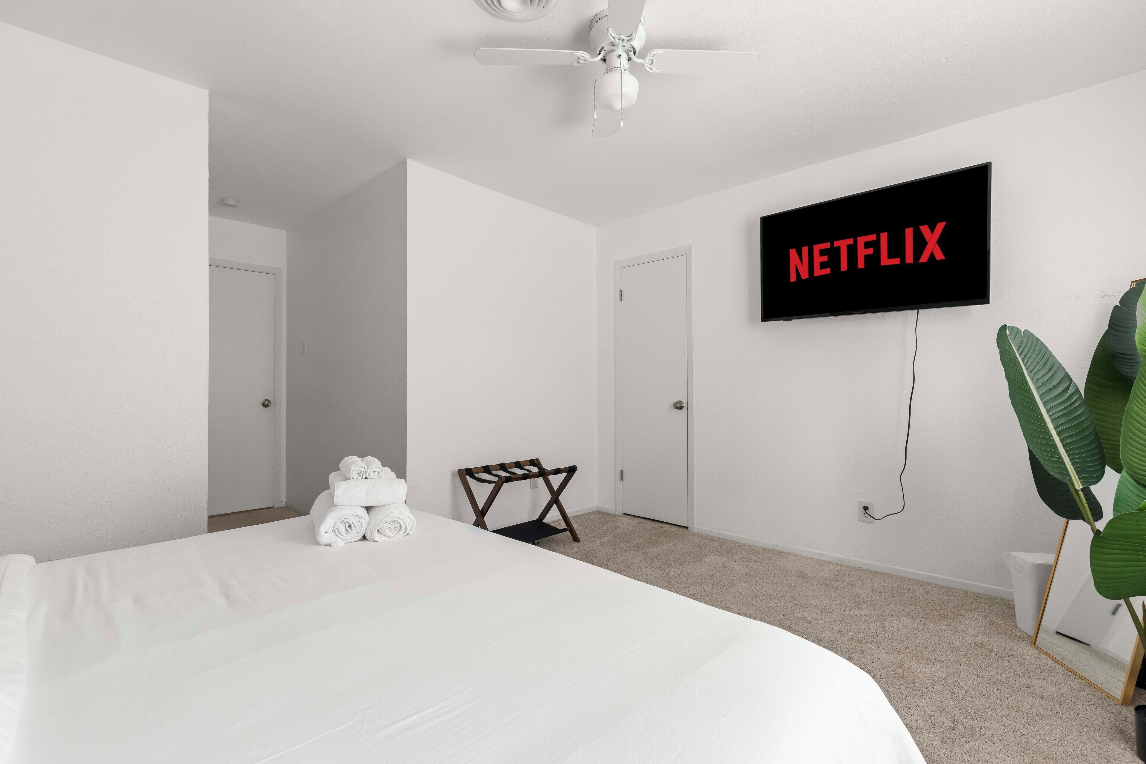 Quarto de hotel com uma cama grande, um ventilador de teto, uma TV na parede exibindo “NETFLIX”, uma cadeira de madeira pequena, uma planta e uma porta.