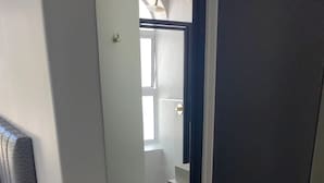 Corredor estreito que dá para um banheiro com chuveiro e uma janela.
