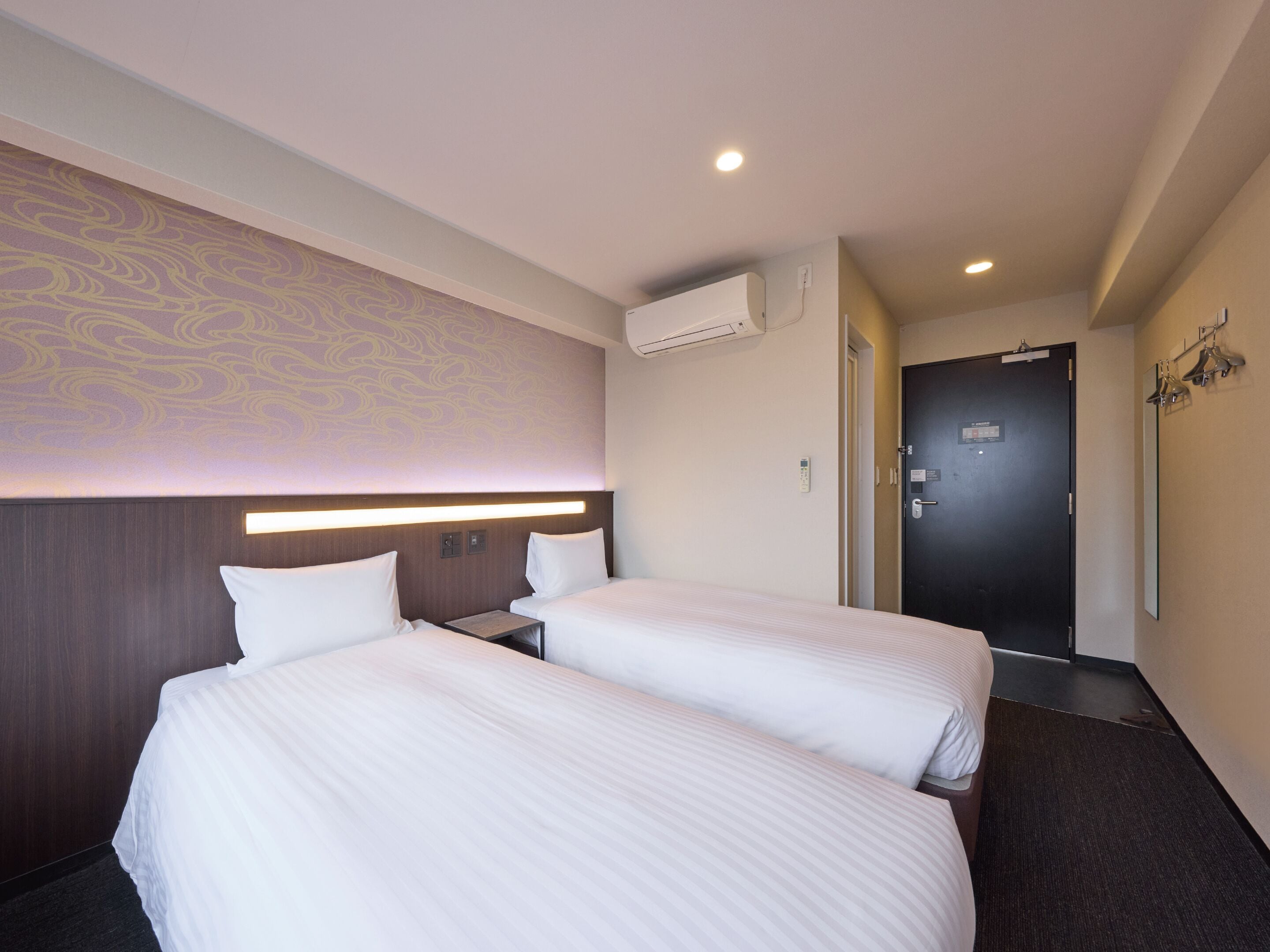 Ein Hotelzimmer mit zwei Betten, einem Kopfteil mit integrierter Beleuchtung, einer Wandklimaanlage und einer Tür mit Spiegel und Lampen.