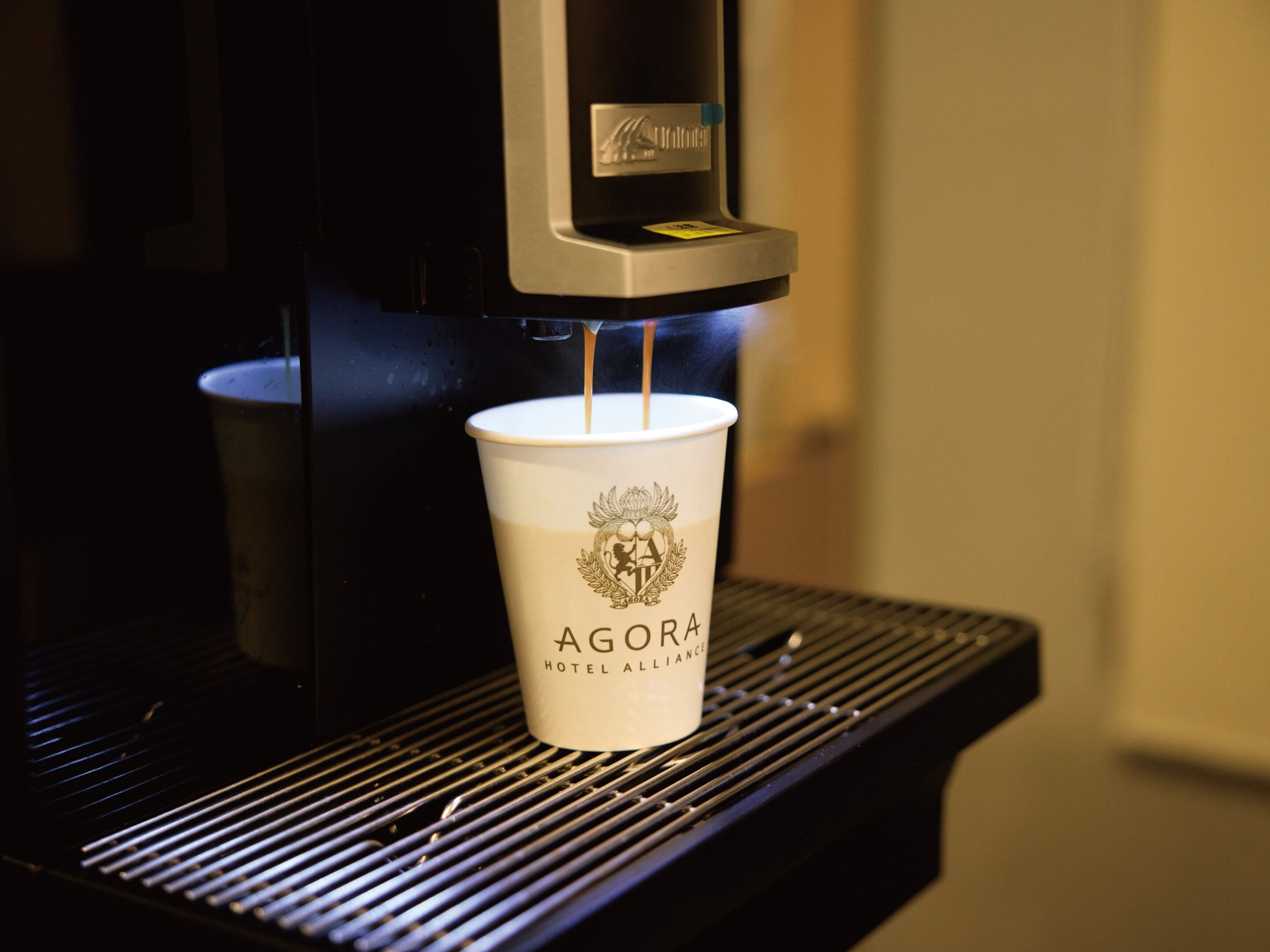Una taza de papel con el logotipo 'AGORA HOTEL ALLIANCE' sobre una máquina de café.