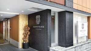 A modern building entrance with a black tiled facade, a logo above the name 'Agora Place,' and a potted plant on the left.