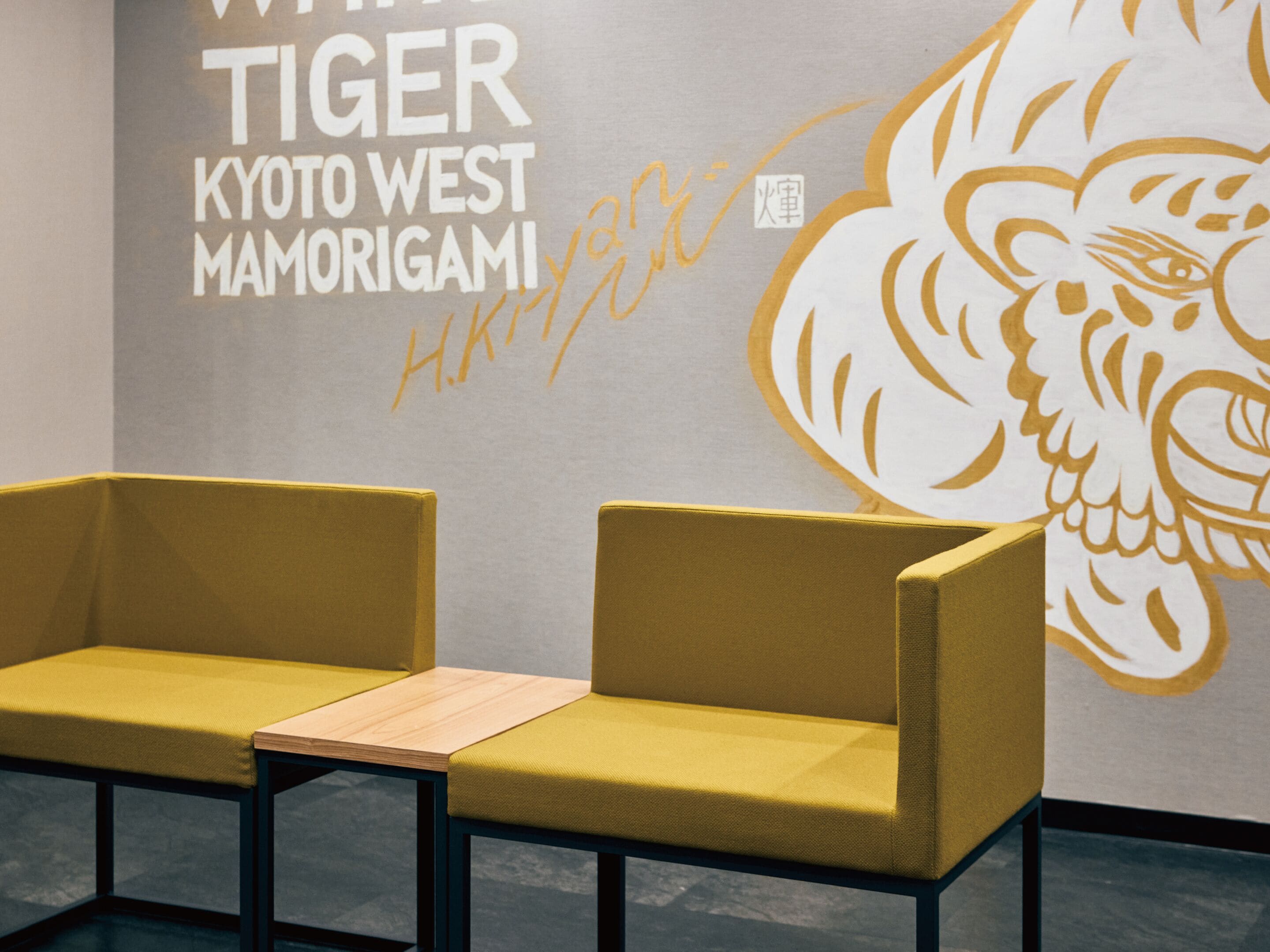 Dos sillas amarillas con asientos de madera, una pared con un mural de tigre, y el texto 'TIGER KYOTO WEST MAMORIGAMI'.