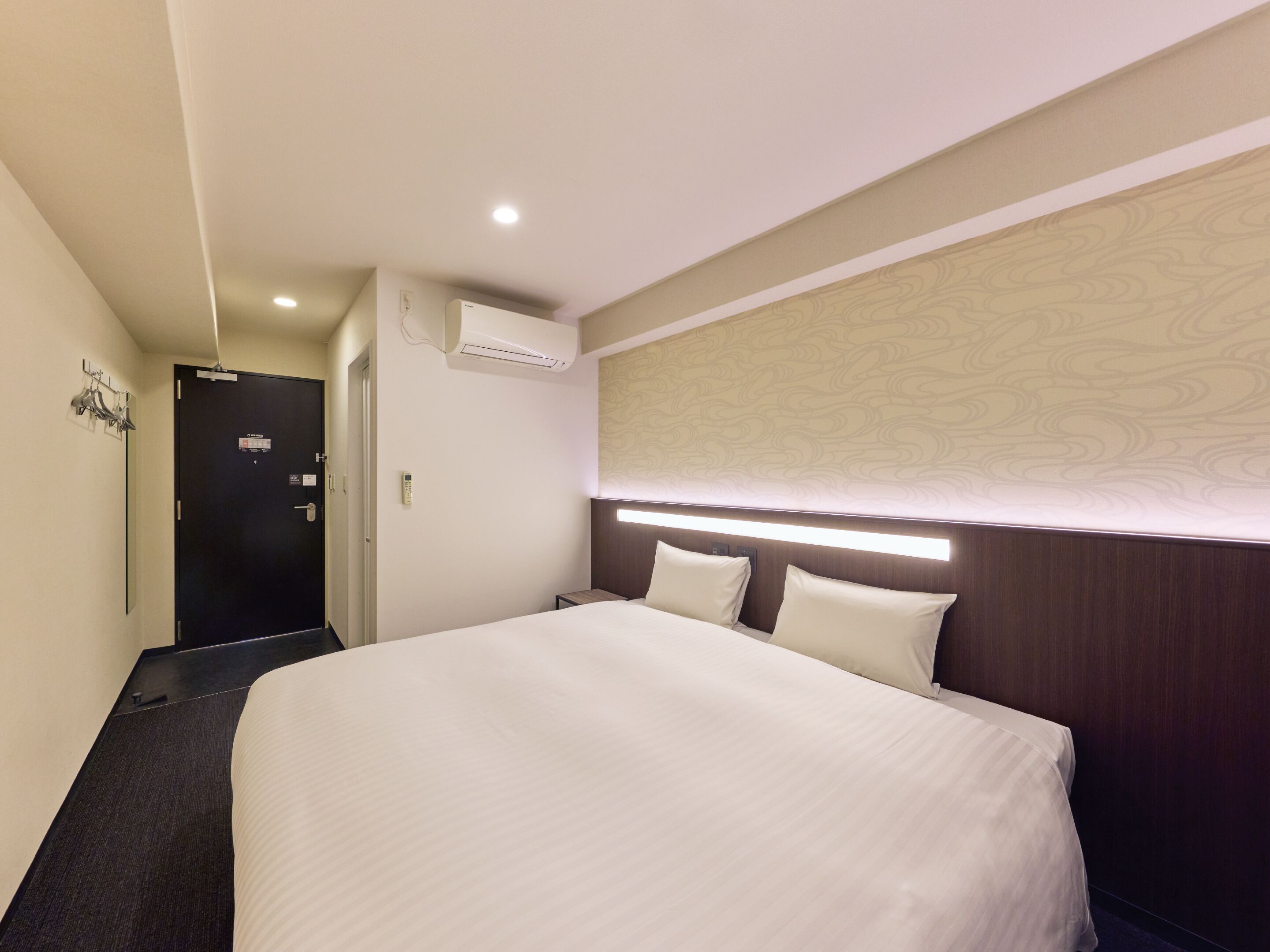Una habitación de hotel con una cama, un equipo de aire acondicionado, una puerta y una lámpara fijada a la pared.