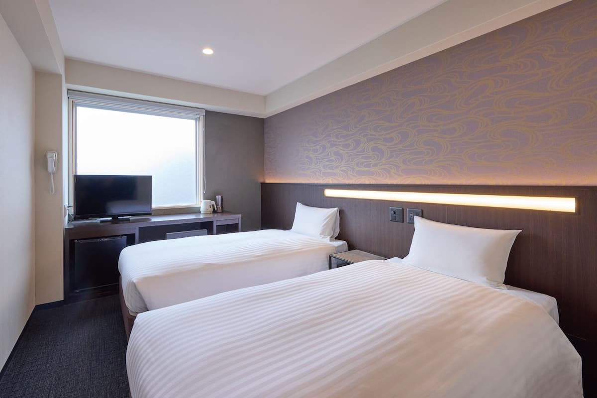 Habitación de hotel con dos camas, un escritorio y un televisor, y una ventana.