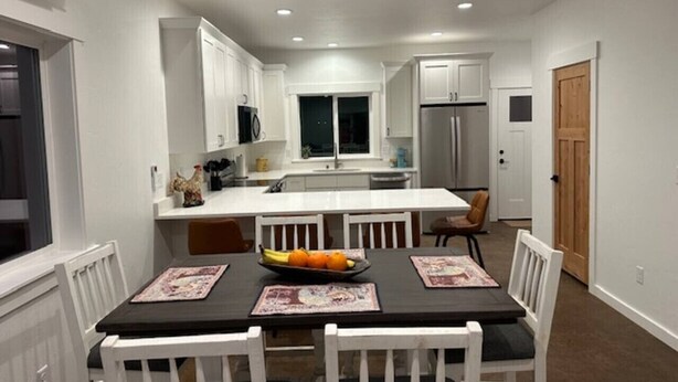 A modern kitchen with white cabinetry, a central island, and a dining area with a black table and white chairs. - The Dam Lakehouse B, Fishing & Boating Basecamp (Grand Coulee)