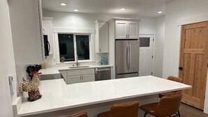 A modern kitchen with white cabinets, a large island with bar stools, a stainless steel refrigerator, and a wooden door. - The Dam Lakehouse B, Fishing & Boating Basecamp (Grand Coulee)
