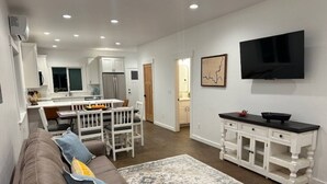A modern living room with a brown sofa, a dining area with a white table and chairs, a kitchen with white cabinets, and a flat-screen TV mounted on the wall. - The Dam Lakehouse B, Fishing & Boating Basecamp (Grand Coulee)