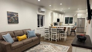 A modern living room with a brown sofa, a dining table with chairs, a kitchen area with white cabinets and stainless steel appliances, and a painting on the wall. - The Dam Lakehouse B, Fishing & Boating Basecamp (Grand Coulee)