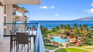 A balcony with a table and chairs, overlooking a pool area with palm trees and a view of the ocean. - Maui Resort Rentals: Honua Kai - 3 Total Bedrooms, Stunning 6th Floor Ocean AND West Maui Mountain Views! (Lahaina)