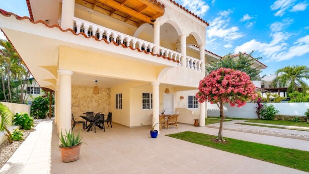 Une maison à deux étages dotée d’un patio couvert, de mobilier d’extérieur et d’un jardin agrémenté d’un arbre en fleurs.