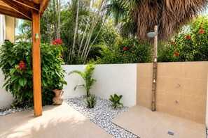 Een overdekt buitengebied met een betegelde vloer, een houten pergola en verschillende planten, waaronder een grote struik met rode bloemen.