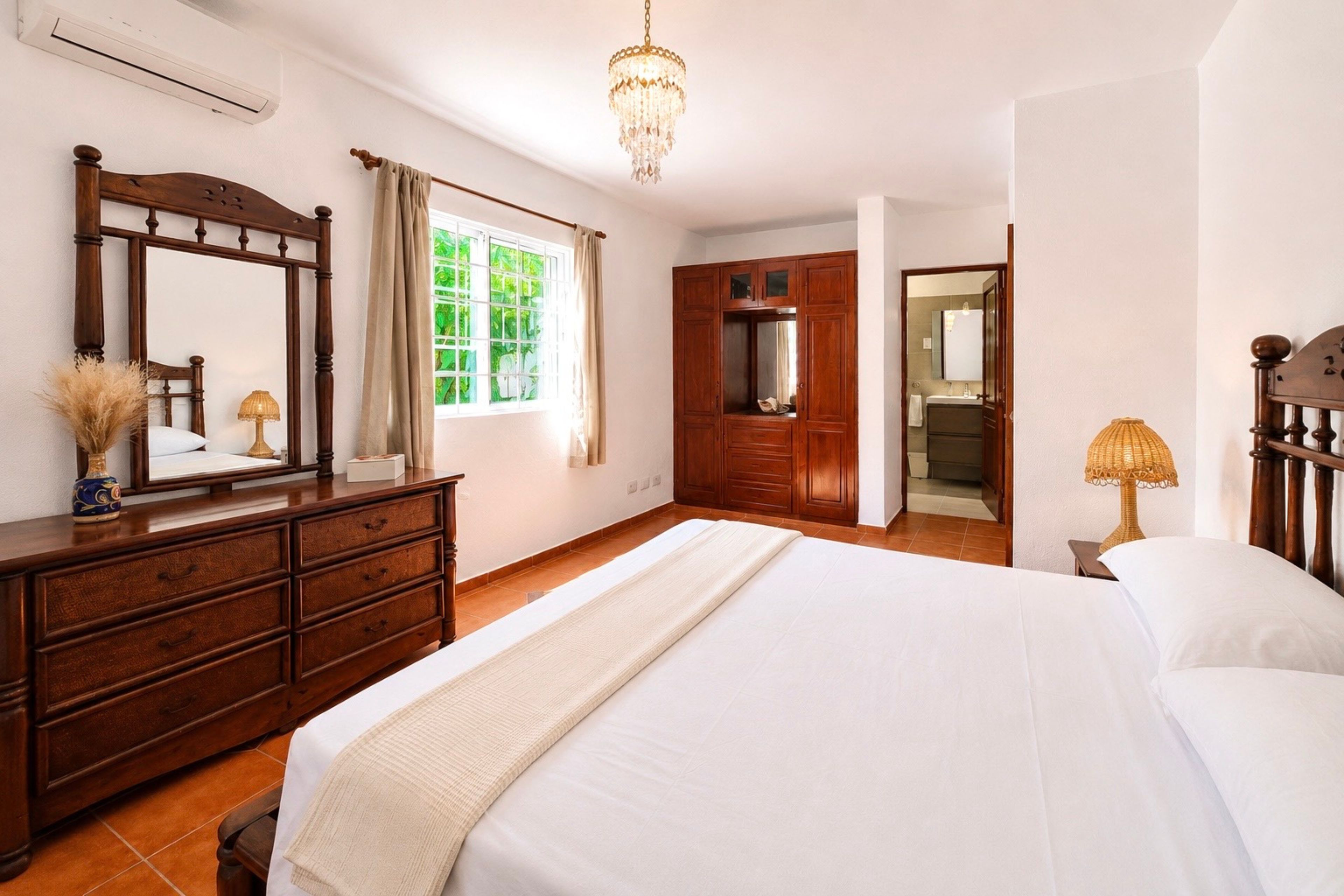 A bedroom with a large bed, a dresser, a mirror, a chandelier, and a view of the bathroom.
