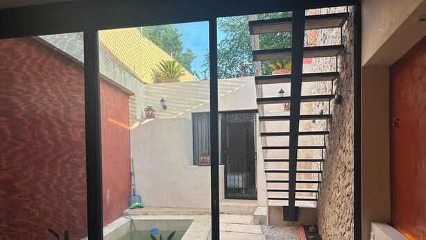 A view through a glass door into a courtyard with a small pool, potted plants, and a staircase leading to a balcony. - Casa Bélanger (Mérida)