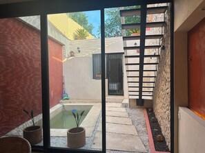 Blick durch eine Glastür in einen Innenhof mit einem kleinen Pool, Kübelpflanzen und einer Treppe, die zu einem Balkon führt.