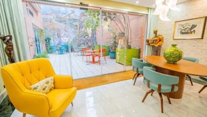 A bright yellow armchair with a cushion, a wooden dining table with chairs, and a view of an outdoor patio with plants and chairs. - Hotel Sancho (Madrid)