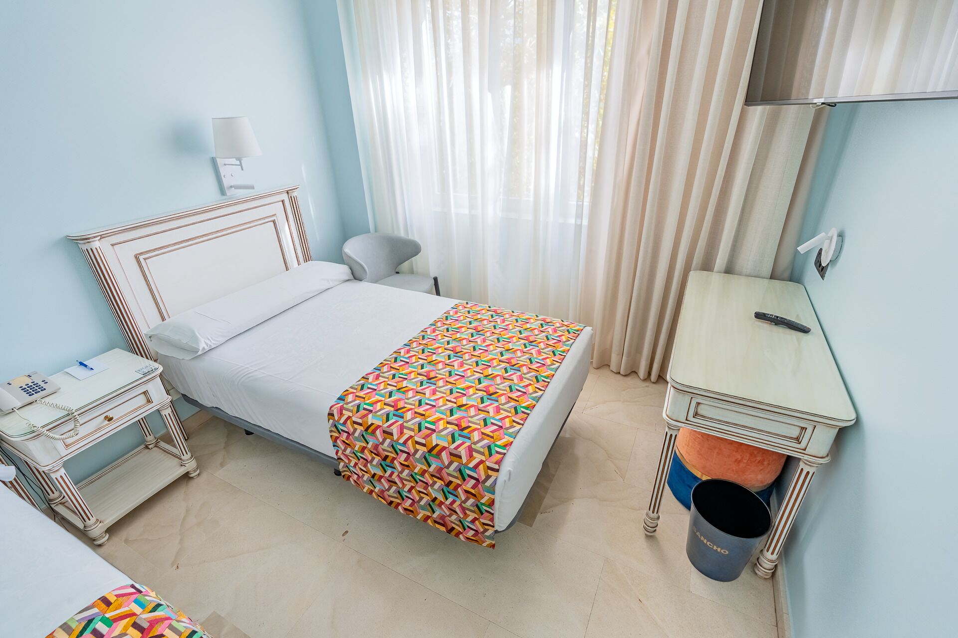 Une chambre d’hôtel comprenant un lit, une table de chevet avec une lampe, une petite table avec une télécommande et une poubelle.