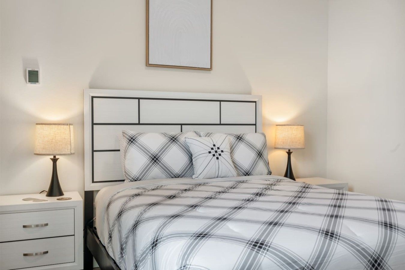 Ein ordentlich bezogenes Bett mit einer Karo-Decke und zwei Kissen, an beiden Seiten mit Nachttischen mit Lampen, in einem Zimmer mit schlichtem, klarem Design.