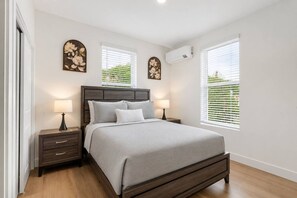 Une chambre à coucher comprenant un lit, des tables de chevet, un climatiseur mural et des fenêtres équipées de stores.