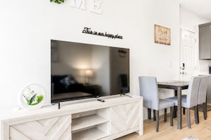 Een moderne woonkamer met een grote flatscreen televisie aan een witte muur, een eethoek met grijze stoelen en een klein bijzettafeltje met een decoratief bord.