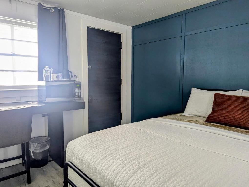 A bedroom with a bed, a desk, a chair, a trash bin, and a door.