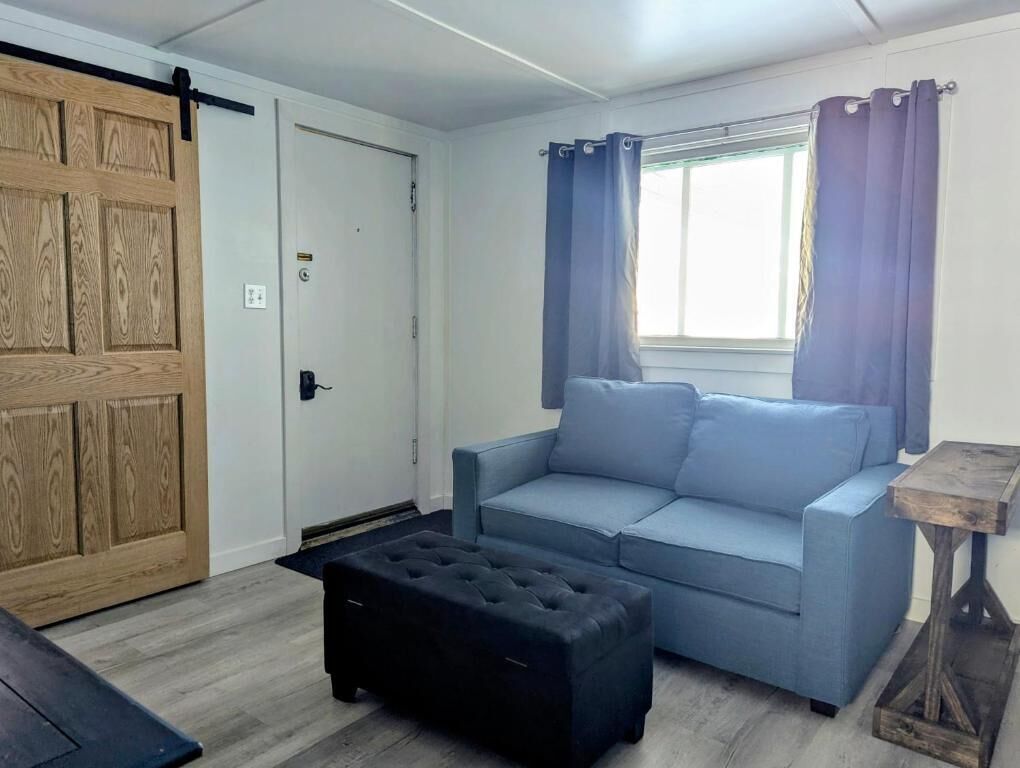 A room with a light blue sofa, a black ottoman, a wooden door, and a window with curtains.