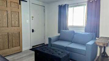 Una habitación con un sofá azul claro, un reposapiés negro, una puerta de madera y una ventana con cortinas.