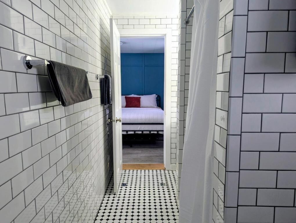 A narrow hallway with white tiled walls and floor, leading to a room with a bed and blue wall.