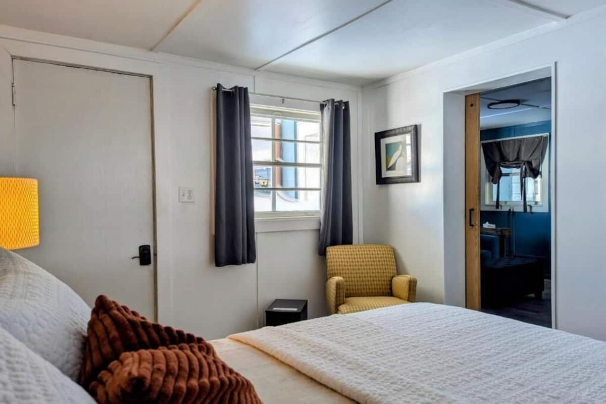 Ett hotellrum med en säng, en stol, ett litet bord, ett fönster med gardiner och en dörr som leder till ett annat rum.