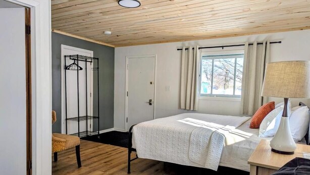 Une chambre à coucher comprenant un lit, une chaise, une table de chevet avec une lampe, une fenêtre avec des rideaux et un porte-manteau.