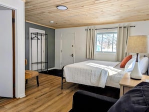 A bedroom with a bed, a chair, a bedside table with a lamp, a window with curtains, and a coat rack. - Scotty's Motel (Munising)